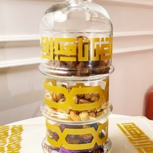 Stacked glass jars with gold geometric designs hold dates, pistachios, and wrapped candies on a decorative, patterned tray. Elegant and festive.