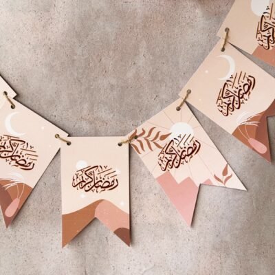 Festive hanging flags with Arabic calligraphy on a textured wall. The design features crescent moons and abstract landscapes. Pink flowers add a touch of color.