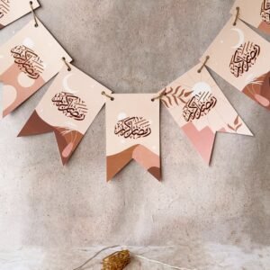 Decorative banner with Arabic calligraphy on beige and brown pennants hangs against a textured wall. Pink flowers and golden lantern string lights enhance the festive tone.