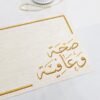 A textured paper with gold Arabic calligraphy is shown close up, displaying intricate letters. To the upper left, a string of pearls and a decorative white lantern are placed on a white surface, conveying an elegant and serene atmosphere.