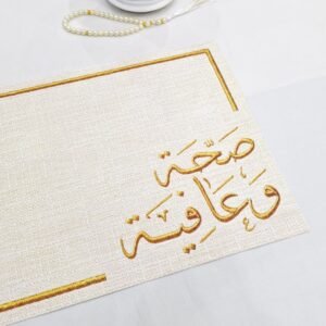 A textured paper with gold Arabic calligraphy is shown close up, displaying intricate letters. To the upper left, a string of pearls and a decorative white lantern are placed on a white surface, conveying an elegant and serene atmosphere.