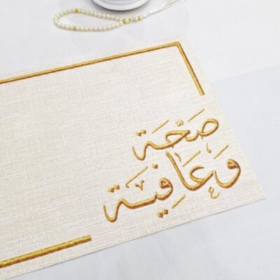 A textured paper with gold Arabic calligraphy is shown close up, displaying intricate letters. To the upper left, a string of pearls and a decorative white lantern are placed on a white surface, conveying an elegant and serene atmosphere.