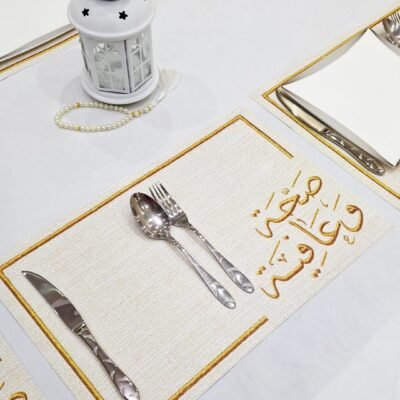 A white tablecloth with square placemats displaying Arabic calligraphy in gold. Cutlery is neatly arranged, next to a decorative lantern and pearl beads.