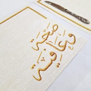 Elegant table setting with a cream-colored placemat featuring gold Arabic calligraphy. Nearby are a shiny knife, fork, and prayer beads, creating a sophisticated and serene ambiance.