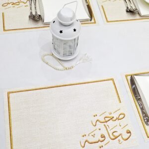 Elegant table setting with white plates and cutlery on gold-accented placemats featuring Arabic text. A white lantern and pearl beads add decor.