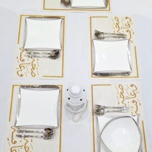 Table set for four with square white plates, shiny silverware, and a decorative lantern centerpiece on ornate gold-patterned placemats. Clean and elegant.