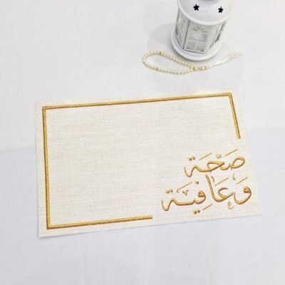 White table with a small white lantern, a string of pearls, and a decorative card featuring gold Arabic calligraphy framed in gold. Peaceful ambiance.
