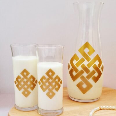 A glass carafe and two glasses filled with milk, featuring gold geometric patterns, rest on a wooden board. A pearl bracelet lies nearby, creating a serene and elegant atmosphere.