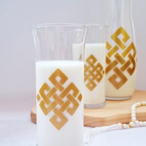 A glass and carafe filled with milk sit on a wooden tray. Both feature a gold geometric design. The scene conveys elegance and simplicity.