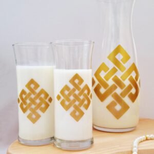 Two glasses and a carafe of milk with golden geometric patterns on a wooden tray, set against a neutral background. A pearl bracelet lies beside them.