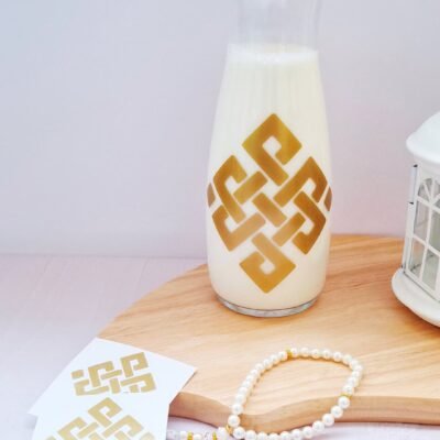 A glass carafe of milk with a gold geometric design sits on a wooden board, accompanied by a pearl bracelet and matching patterned cards, evoking elegance.