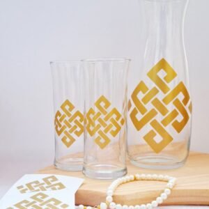Glass carafe and two tumblers with golden geometric patterns on a wooden board. A pearl bracelet and matching patterned cards add elegance.