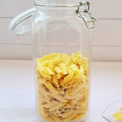 A clear glass jar with a metal clasp lid is filled with uncooked penne pasta, set on a white surface. A few pasta pieces lie beside it, creating a simple, organized scene.