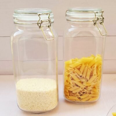 Two glass jars with metal clasps on a light surface. The left jar is half-filled with rice, and the right jar is half-filled with penne pasta.