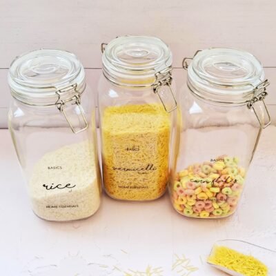 Three glass jars with metal clasps on a light wooden surface contain rice, vermicelli, and colorful cornflakes. A small dish with spilled vermicelli lies nearby.