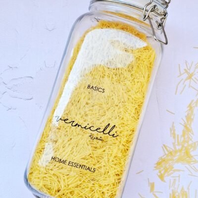 Clear glass jar filled with yellow vermicelli pasta, labeled “Basics Vermicelli” and “Home Essentials.” Some pasta spills out onto a white surface.