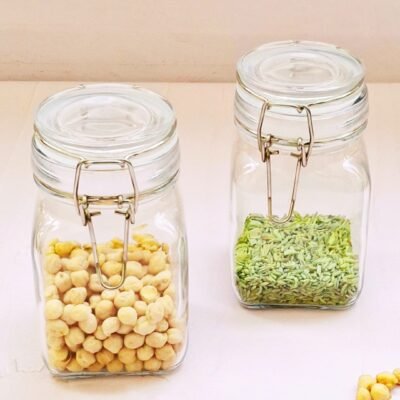 Two glass jars with clip lids on a light surface, one filled with chickpeas and the other with green fennel seeds. Some chickpeas are scattered nearby.