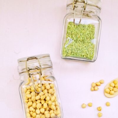 Two glass jars with metal clasps on a light surface; one contains chickpeas, the other fennel seeds. A few chickpeas are scattered around.