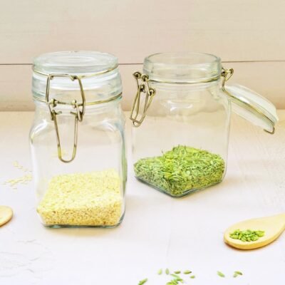 Two glass jars on a light wooden surface; one contains white sesame seeds, the other has green fennel seeds. Loose seeds and wooden spoons surround them.