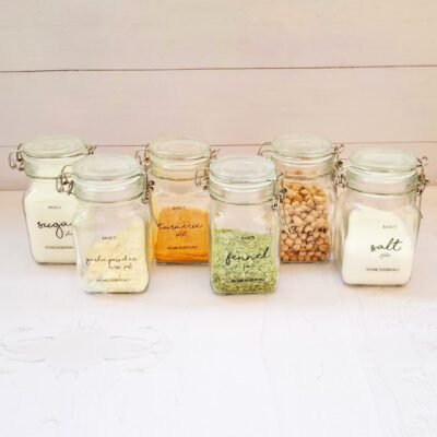 Six glass jars with metal clasps labeled with contents: sugar, garlic powder, turmeric, fennel, chickpeas, and salt, arranged on a light wooden surface.