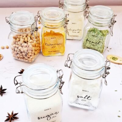 Six labeled glass jars, including chickpeas, turmeric, garlic powder, fennel, sugar, and salt, arranged on a bright surface with star anise.
