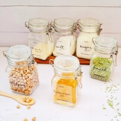 Six glass jars filled with various kitchen essentials, including chickpeas, turmeric, and fennel, are displayed neatly against a white background.