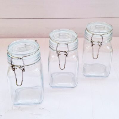 Three transparent glass jars with metal clasps are arranged on a white wooden surface. The jars are empty, conveying a clean, minimalist look.