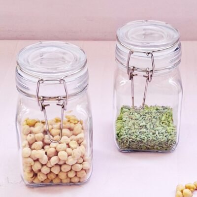 Two glass jars with clip lids on a light surface, one filled with chickpeas and the other with green fennel seeds. Some chickpeas are scattered nearby.