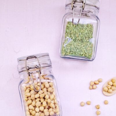 Two glass jars with metal clasps on a light surface; one contains chickpeas, the other fennel seeds. A few chickpeas are scattered around.