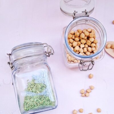 Two glass jars on a light surface. One jar, lying down, contains green fennel seeds. The other, upright, is filled with chickpeas, some scattered. Calm tone.