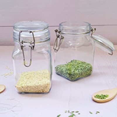 Two glass jars on a light wooden surface; one contains white sesame seeds, the other has green fennel seeds. Loose seeds and wooden spoons surround them.