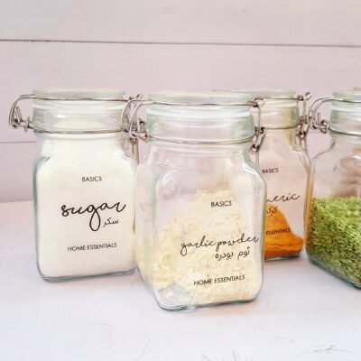 Four labeled glass jars containing sugar, garlic powder, turmeric, and a green herb are placed in a row on a white surface, creating a neat, organized appearance.