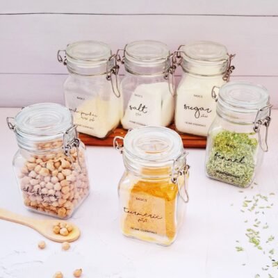 Six glass jars filled with various kitchen essentials, including chickpeas, turmeric, and fennel, are displayed neatly against a white background.