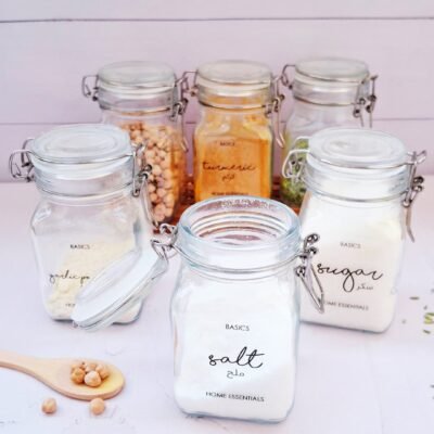 Six glass jars with metal clasps are filled with various kitchen staples, such as salt, sugar, and spices, labeled neatly. The setting is clean and organized.