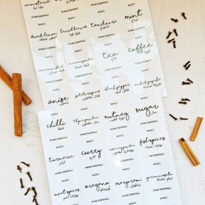 Sheets of spice and herb labels in English and Arabic with flowing script font. Cloves and cinnamon sticks are scattered around on a light surface.
