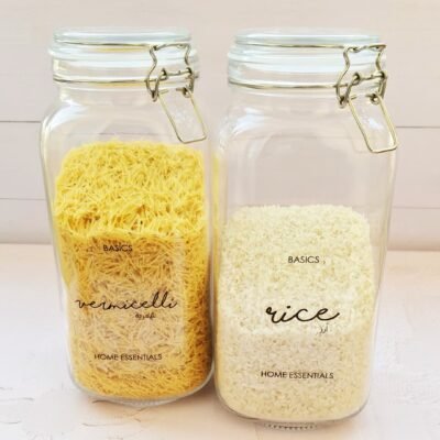 Two glass jars with metal clasps sit on a light surface. The left jar is filled with yellow vermicelli, and the right jar contains white rice. Both jars are labeled 