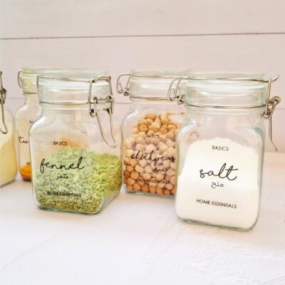 Five glass jars with metal clasps are filled with spices and legumes, including fennel, chickpeas, and salt, labeled in elegant black script.