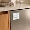Stainless steel dishwasher with a magnet labeled "CLEAN" and "DIRTY" on the door. Beige dish towels are neatly rolled on the countertop above.