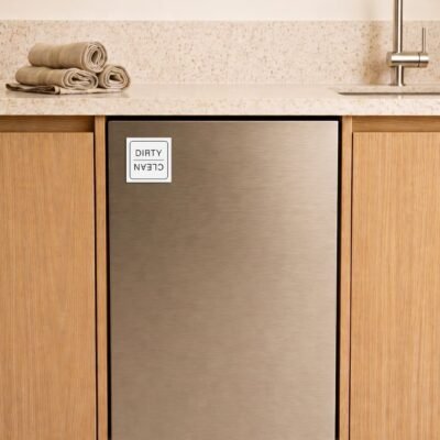 A stainless steel dishwasher is integrated into a wooden kitchen cabinet. A "Dirty/Clean" magnet is on the door. Rolled towels are atop a speckled countertop.