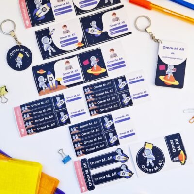 Flat lay of colorful Omar M. Ali stickers, labels, and keychains with playful astronaut designs, plus pens, scissors, and folders.