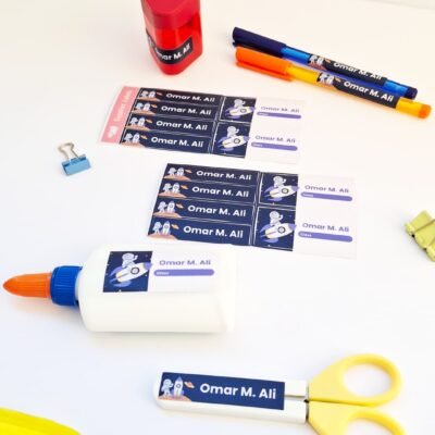 White desk displaying personalized “Omar M. Ali” labels, glue bottle, yellow scissors, colored pens, sharpener, clips, and a folder.
