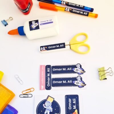 Top-down view of colorful stationery labeled “Omar M. Ali,” including pens, glue, scissors, clips, stickers, and pencil cases.