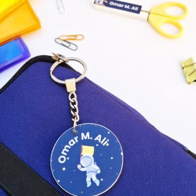 Personalized “Omar M. Ali” astronaut keychain rests on a blue bag, surrounded by colorful stationery, scissors, paper clips, and binder clips.