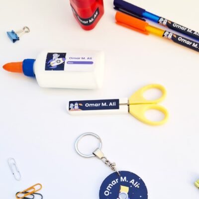 Top-down view of colorful school supplies—glue, markers, scissors, paper clips, sharpener, and an astronaut keychain—on a white background.