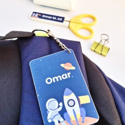 Blue astronaut-themed Omar ID badge hangs from a dark backpack, featuring a rocket and planet design, with scissors, glue, pen, and clip nearby.