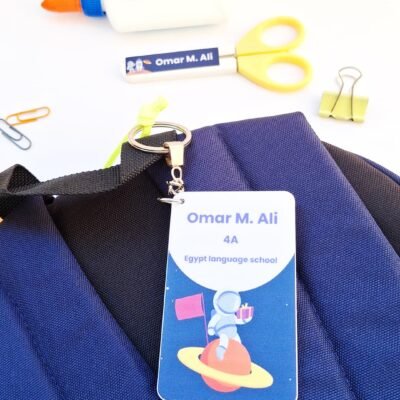 Blue backpack with a space-themed name tag reading “Omar M. Ali, 4A, Egypt language school,” beside scissors, glue, clips, and key ring.