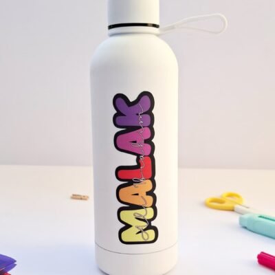 White reusable water bottle with a looped cap and colorful vertical lettering stands on a desk among stationery in a clean, playful scene.