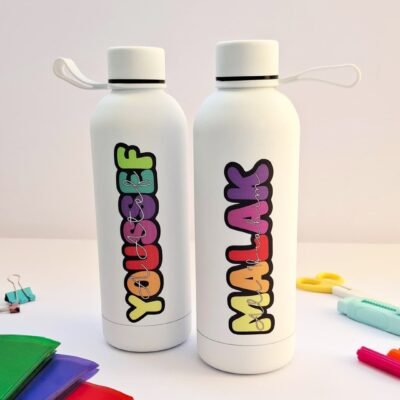 Two white reusable water bottles with colorful vertical lettering stand on a desk, surrounded by bright stationery, creating a playful mood.