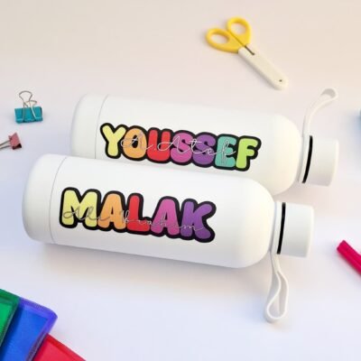 Two white cylindrical bottles with colorful bubble-letter labels reading “YOUSSEF” and “MALAK,” beside scissors, clips, and bright stationery.