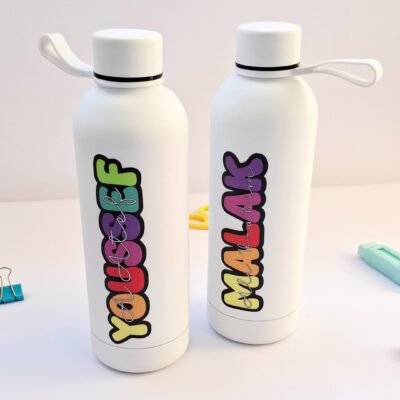 Two white reusable bottles with loop caps and colorful vertical lettering stand on a pastel desk, creating a playful, cheerful mood.